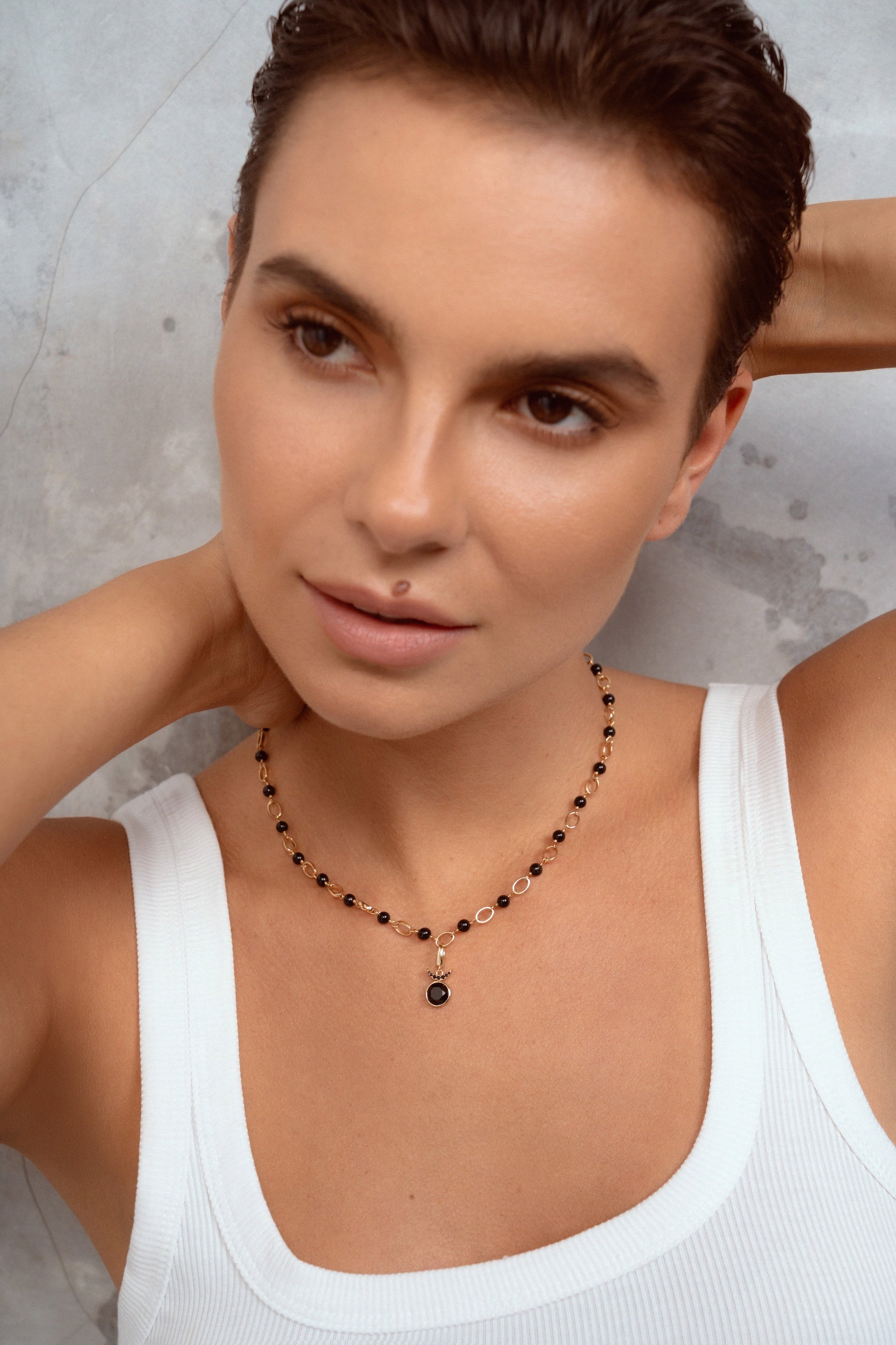 A close-up of a woman with short brown hair wearing a delicate beaded necklace with black and gold beads and a small dark pendant, paired with a white ribbed tank top against a textured gray wall.