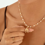 A close-up lifestyle shot of a model wearing a delicate gold chain necklace with small white pearls and a tiny house-shaped pendant, while touching the necklace with her hand.