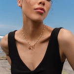Woman wearing a gold and pearl charm necklace featuring a Hecate’s Wheel charm, a triquetra charm, and a crescent moon charm with black and white stones, photographed by the beach.