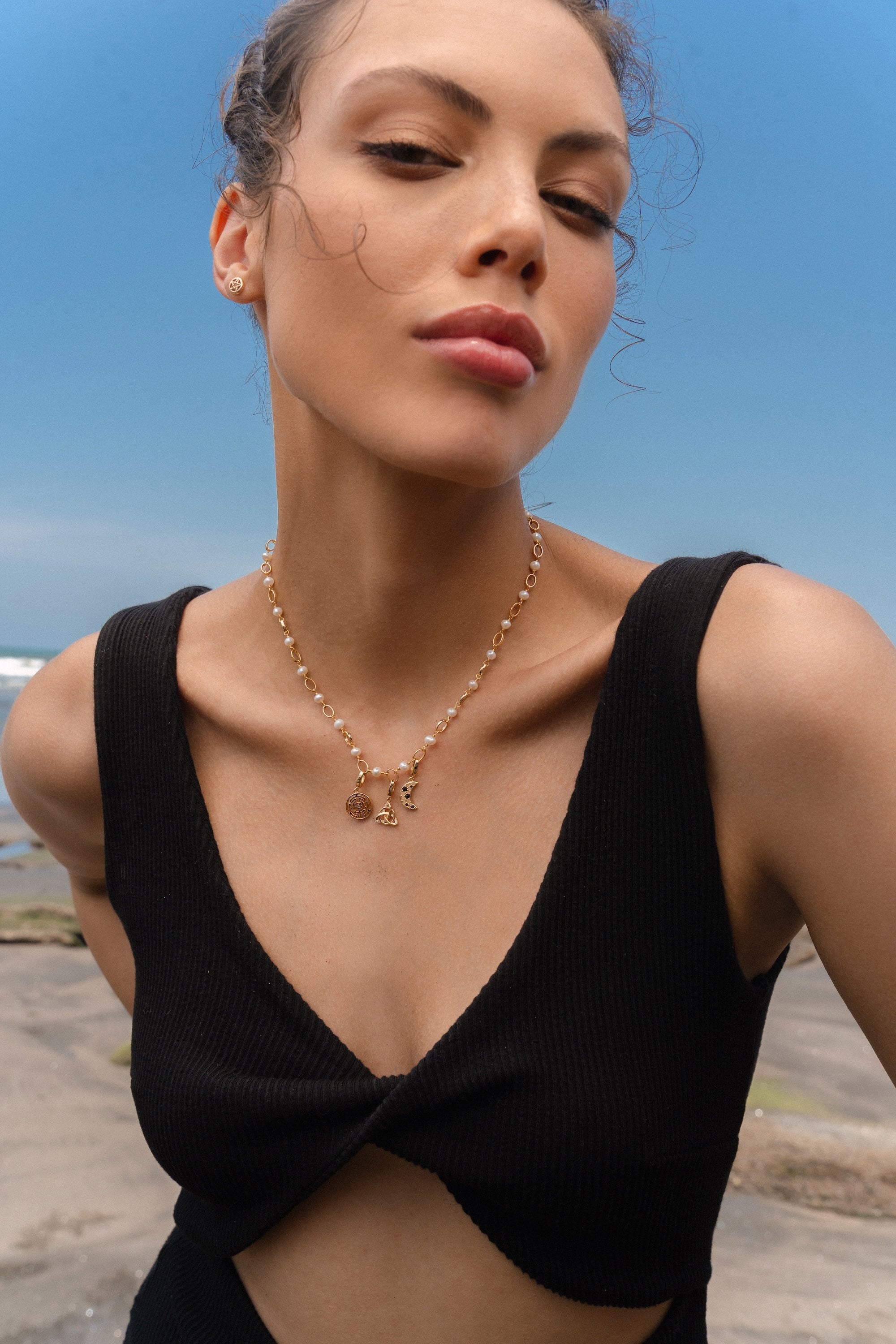 Woman wearing a gold and pearl charm necklace featuring a Hecate’s Wheel charm, a triquetra charm, and a crescent moon charm with black and white stones, photographed by the beach.