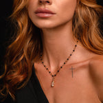 A woman with wavy auburn hair, a black off-shoulder top, and a small cross tattoo on her collarbone wears the Hourglass Charm pendant on a black beaded necklace and poses against a dark background.