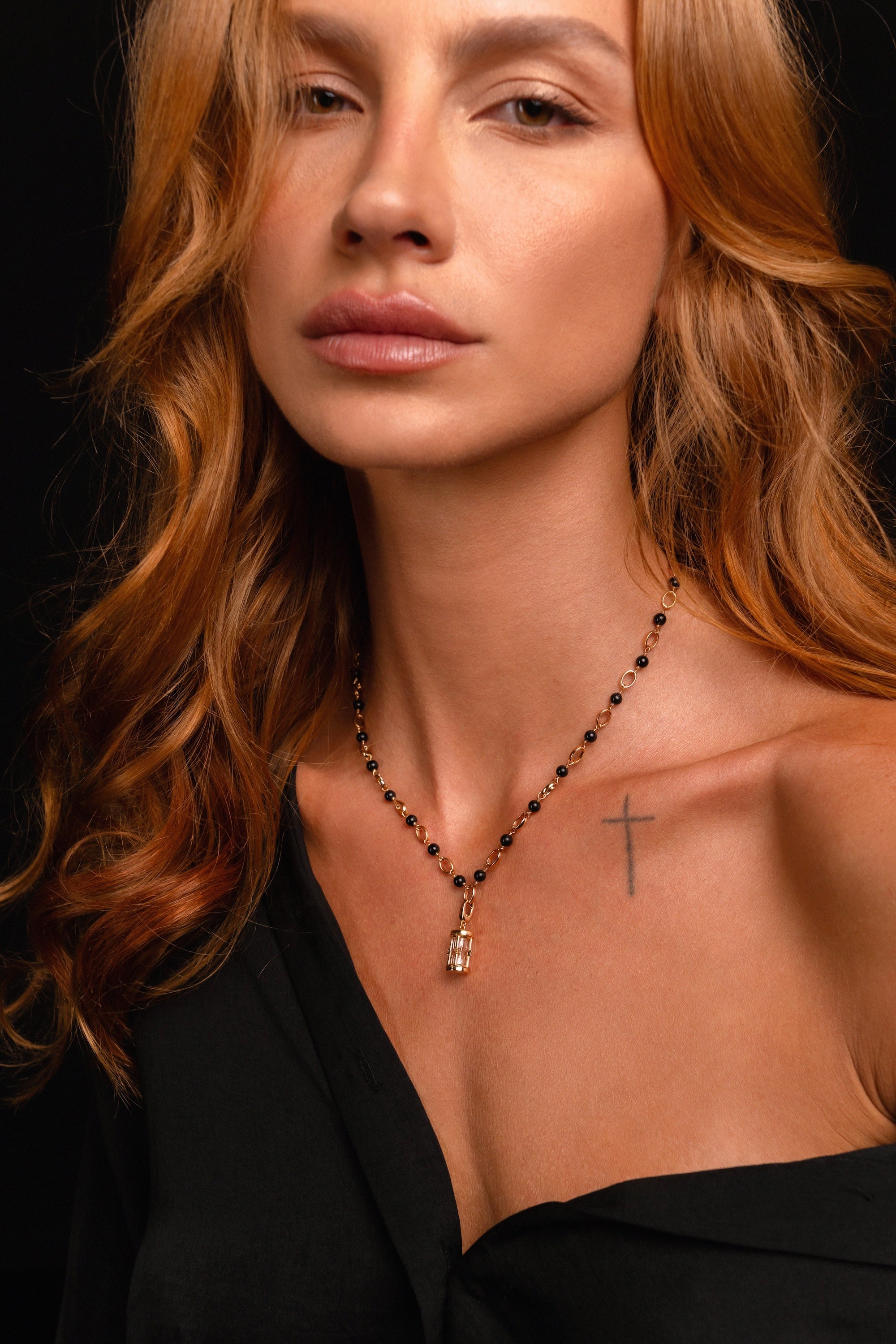 A woman with wavy auburn hair, a black off-shoulder top, and a small cross tattoo on her collarbone wears the Hourglass Charm pendant on a black beaded necklace and poses against a dark background.