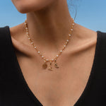 Woman wearing a gold and pearl charm necklace featuring a Hecate’s Wheel charm, a triquetra charm, and a crescent moon charm with black and white stones, photographed by the beach.