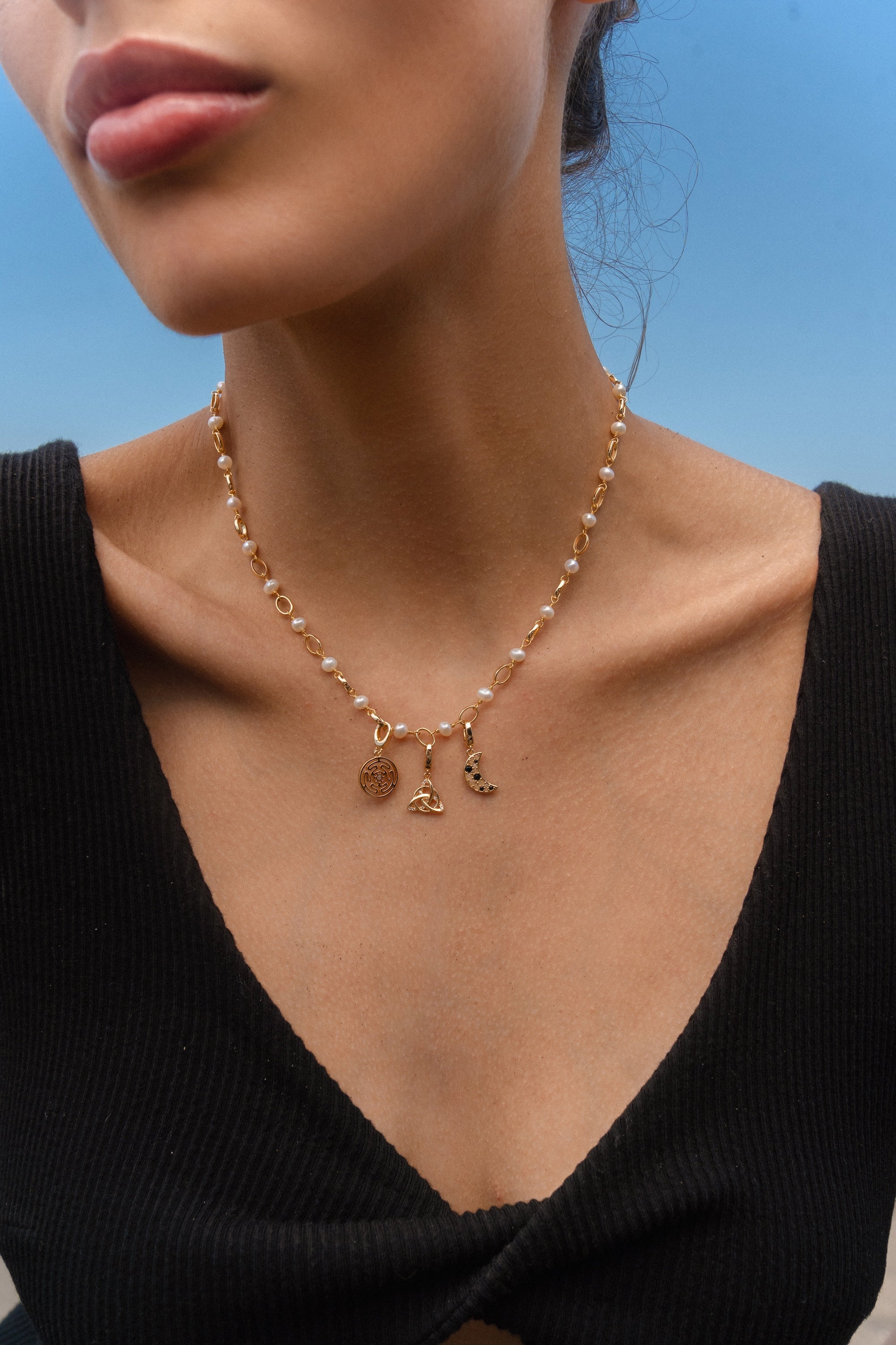 Woman wearing a gold and pearl charm necklace featuring a Hecate’s Wheel charm, a triquetra charm, and a crescent moon charm with black and white stones, photographed by the beach.