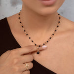 A close-up of a model wearing a delicate necklace with alternating black and gold beads, featuring a small black bead pendant. The model is wearing a black top and a gold ring on her finger.