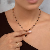 A close-up of a model wearing a delicate necklace with alternating black and gold beads, featuring a small black bead pendant. The model is wearing a black top and a gold ring on her finger.