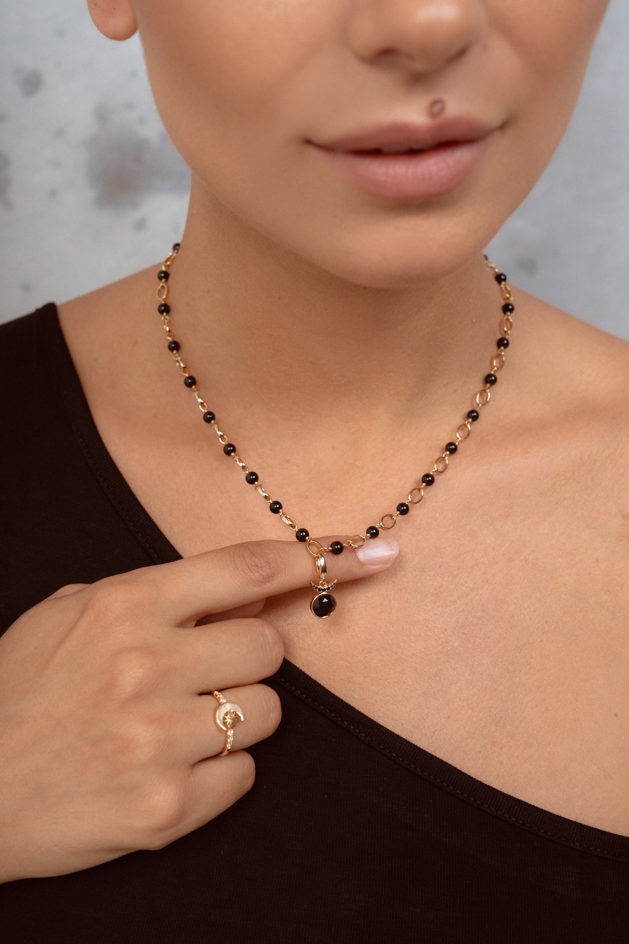 A close-up of a model wearing a delicate necklace with alternating black and gold beads, featuring a small black bead pendant. The model is wearing a black top and a gold ring on her finger.