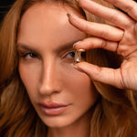A woman with long, light brown hair holds an Hourglass Charm near her eye, gazing into the camera against a dark background.