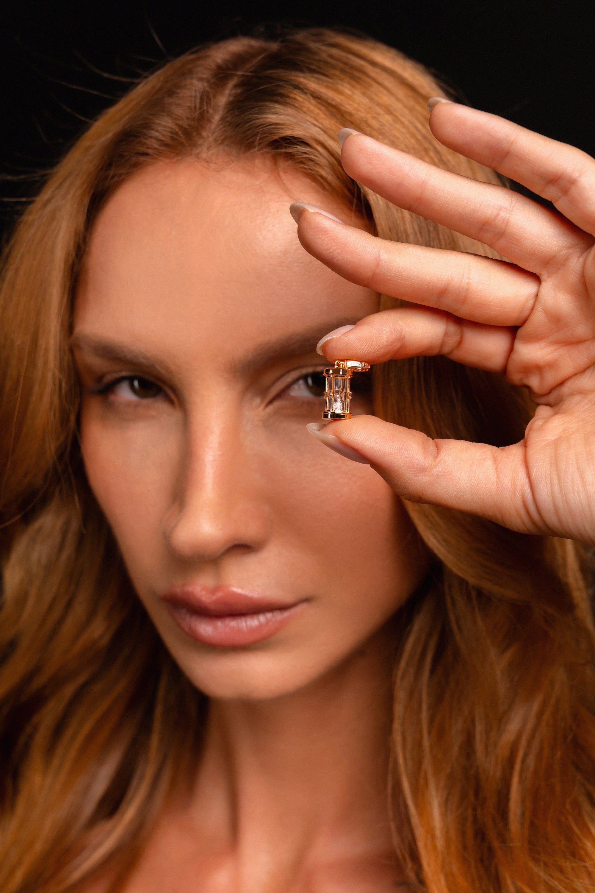 A woman with long, light brown hair holds an Hourglass Charm near her eye, gazing into the camera against a dark background.