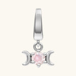 A silver-toned triple moon charm pendant featuring a central round pink gemstone, shown against a solid off-white background.