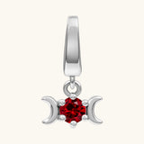 A silver-toned triple moon pendant charm featuring a central faceted red gemstone, set against a solid off-white background.