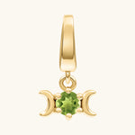 A gold pendant charm featuring a central green peridot gemstone flanked by two crescent moons, shown on a light cream background.