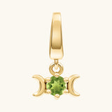 A gold pendant charm featuring a central green peridot gemstone flanked by two crescent moons, shown on a light cream background.