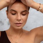 A close-up lifestyle shot of a woman wearing a delicate gold chain necklace with a small black pendant and a matching gold bracelet, set against a textured gray background.