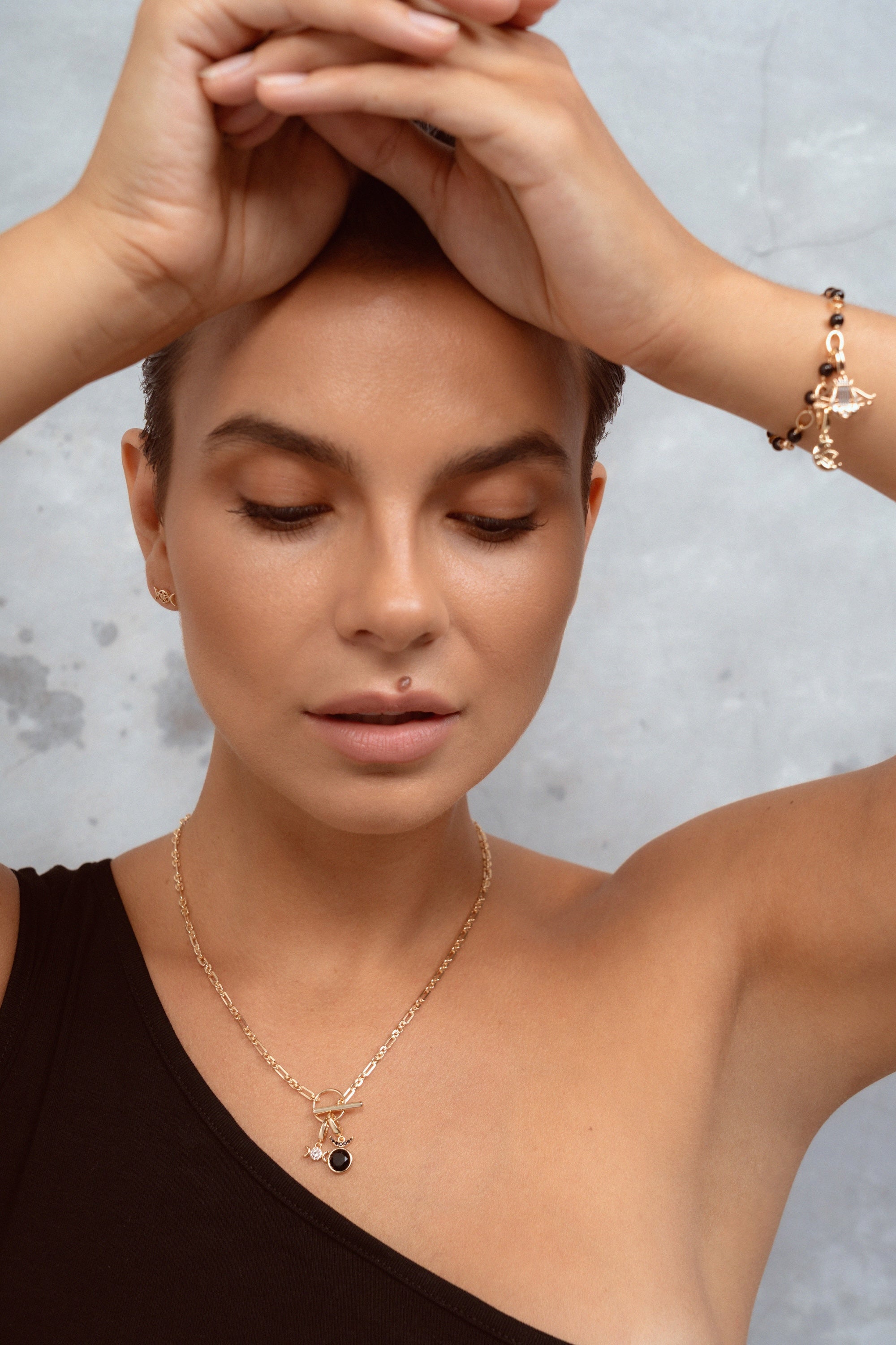A close-up lifestyle shot of a woman wearing a delicate gold chain necklace with a small black pendant and a matching gold bracelet, set against a textured gray background.