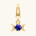 A gold pendant charm featuring a triple moon symbol surrounding a round rich blue gemstone, shown on a light cream background.