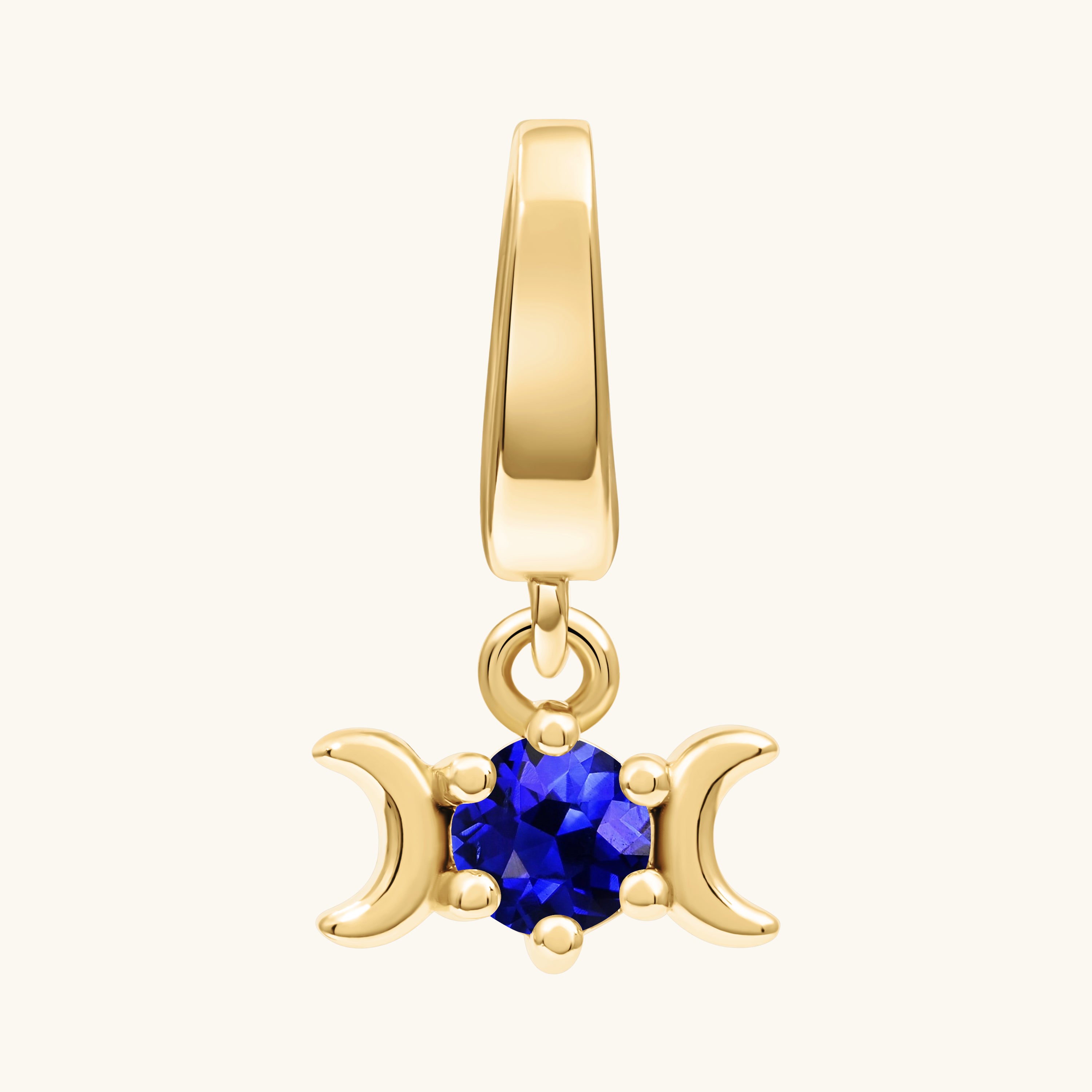 A gold pendant charm featuring a triple moon symbol surrounding a round rich blue gemstone, shown on a light cream background.