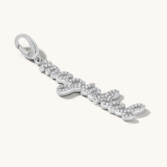 The word magickal is written in elegant cursive script and studded with white topaz stones. It is set in a silver charm pendant against an light cream background. 