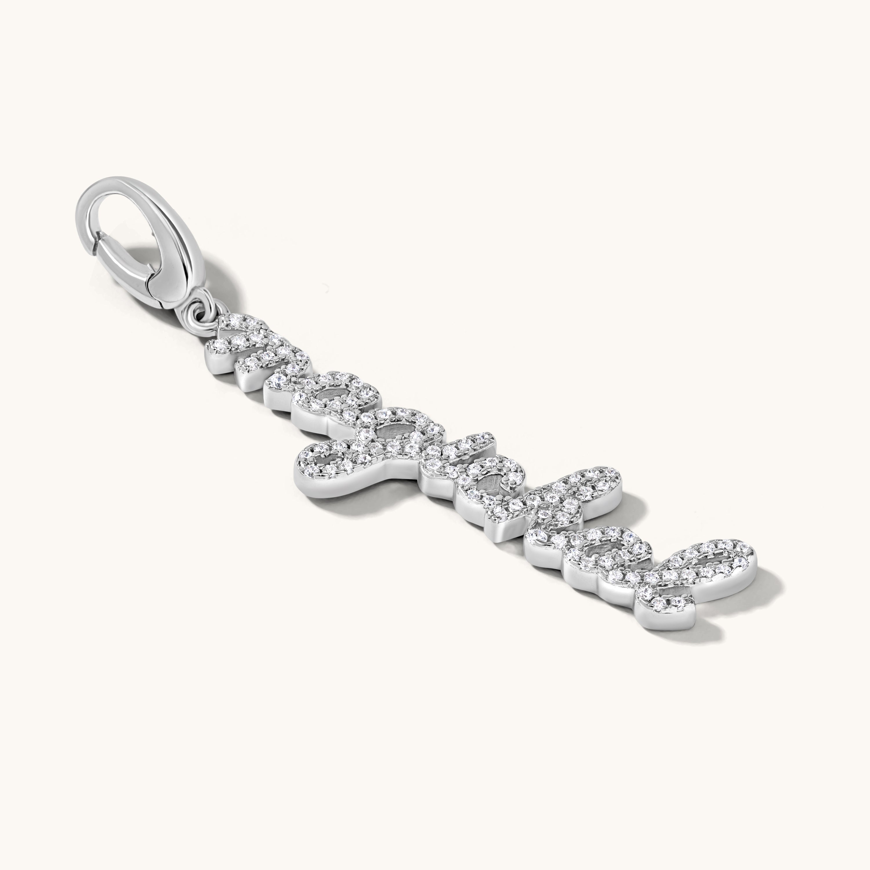 The word magickal is written in elegant cursive script and studded with white topaz stones. It is set in a silver charm pendant against an light cream background. 