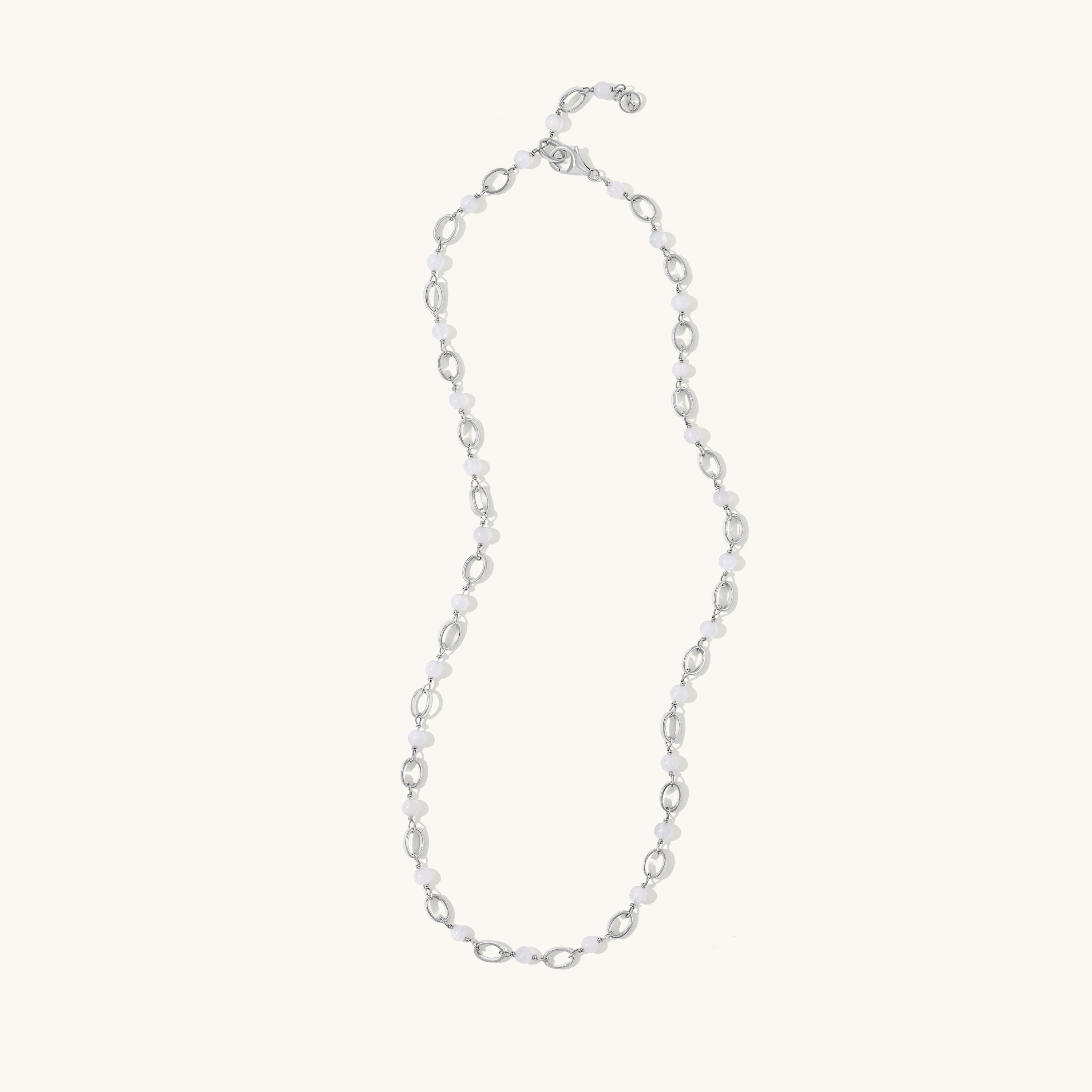 A delicate silver chain necklace with alternating small oval links and tiny clear faceted beads, displayed on a solid off-white background.