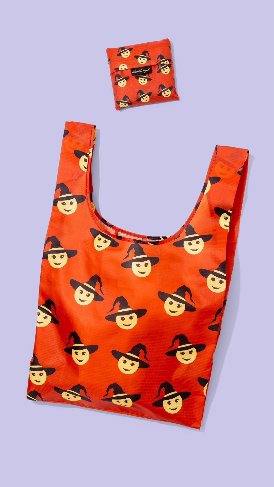 An orange reusable shopping bag with a repeating pattern of emoji faces wearing black witch hats, shown on a light lavender background with a smaller matching bag folded above it.