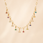A gold chain necklace featuring a series of small dangling Triple Goddess charms, each embellished with a different colored gemstone, displayed against a soft beige background.