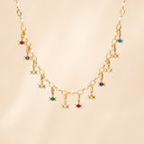 A gold chain necklace featuring a series of small dangling Triple Goddess charms, each embellished with a different colored gemstone, displayed against a soft beige background.