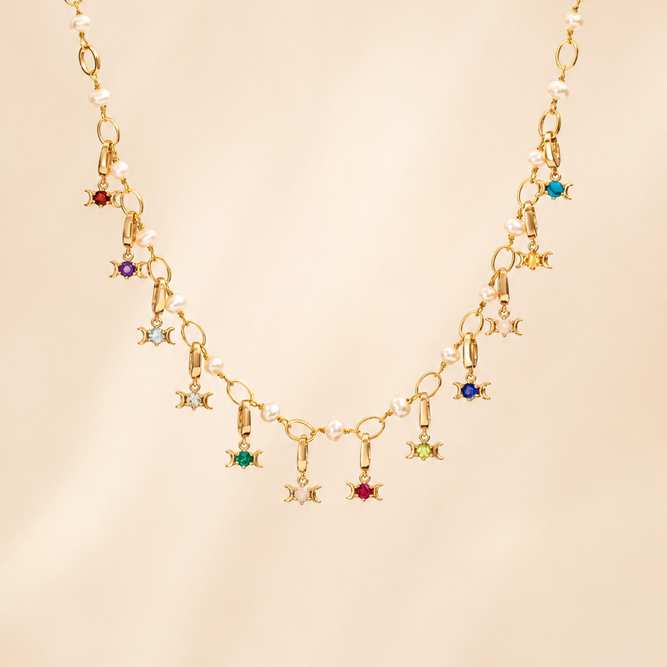 A gold chain necklace featuring a series of small dangling Triple Goddess charms, each embellished with a different colored gemstone, displayed against a soft beige background.