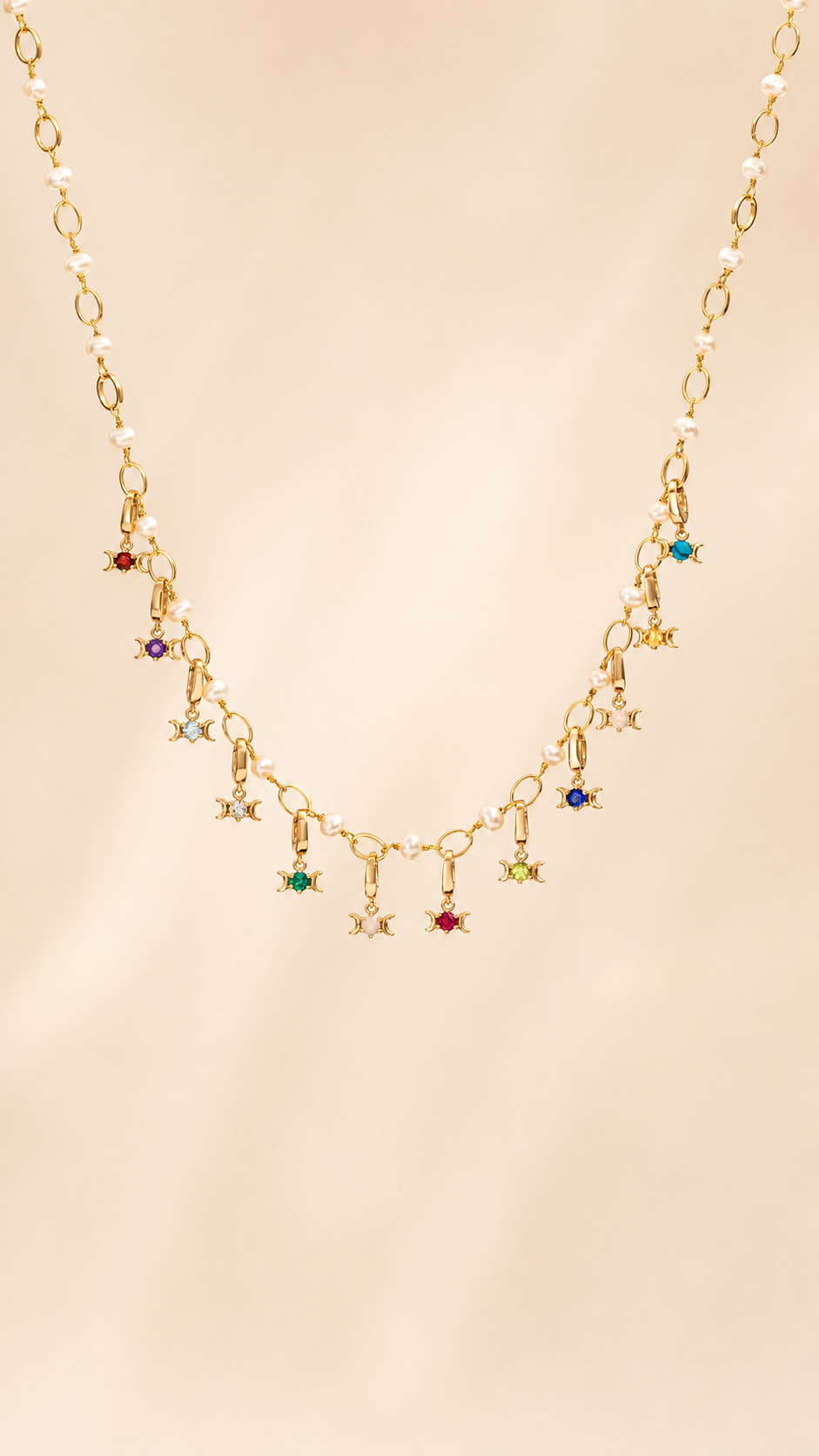 A gold chain necklace featuring a series of small dangling Triple Goddess charms, each embellished with a different colored gemstone, displayed against a soft beige background.