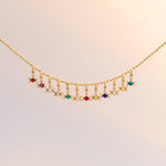 A delicate gold chain necklace featuring a curved row of small dangling charms, including colorful gemstone beads and moon-shaped elements, displayed against a soft, light beige background.