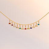 A delicate gold chain necklace featuring a curved row of small dangling charms, including colorful gemstone beads and moon-shaped elements, displayed against a soft, light beige background.