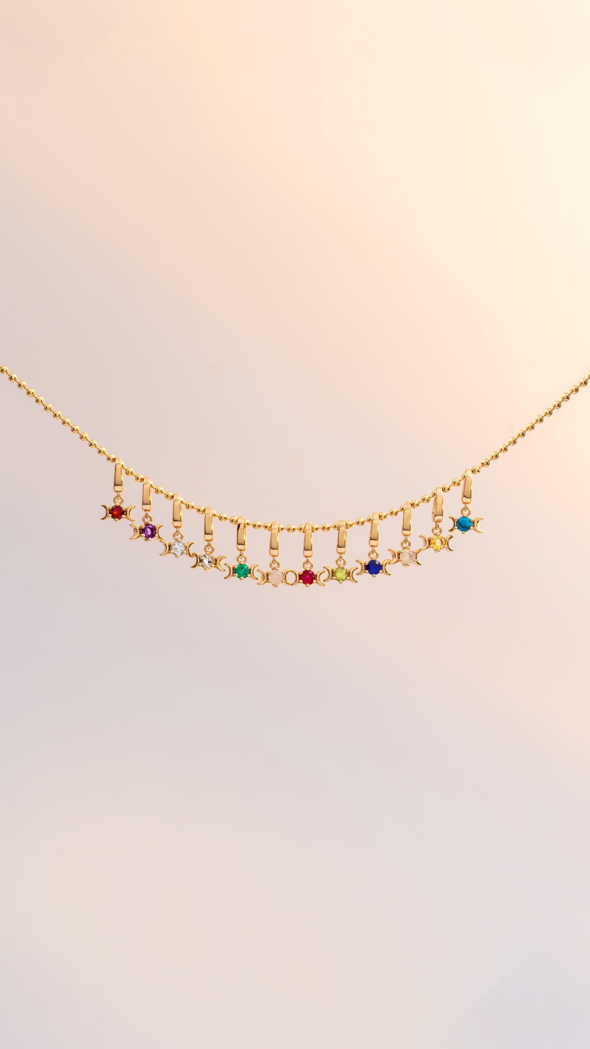 A delicate gold chain necklace featuring a curved row of small dangling charms, including colorful gemstone beads and moon-shaped elements, displayed against a soft, light beige background.
