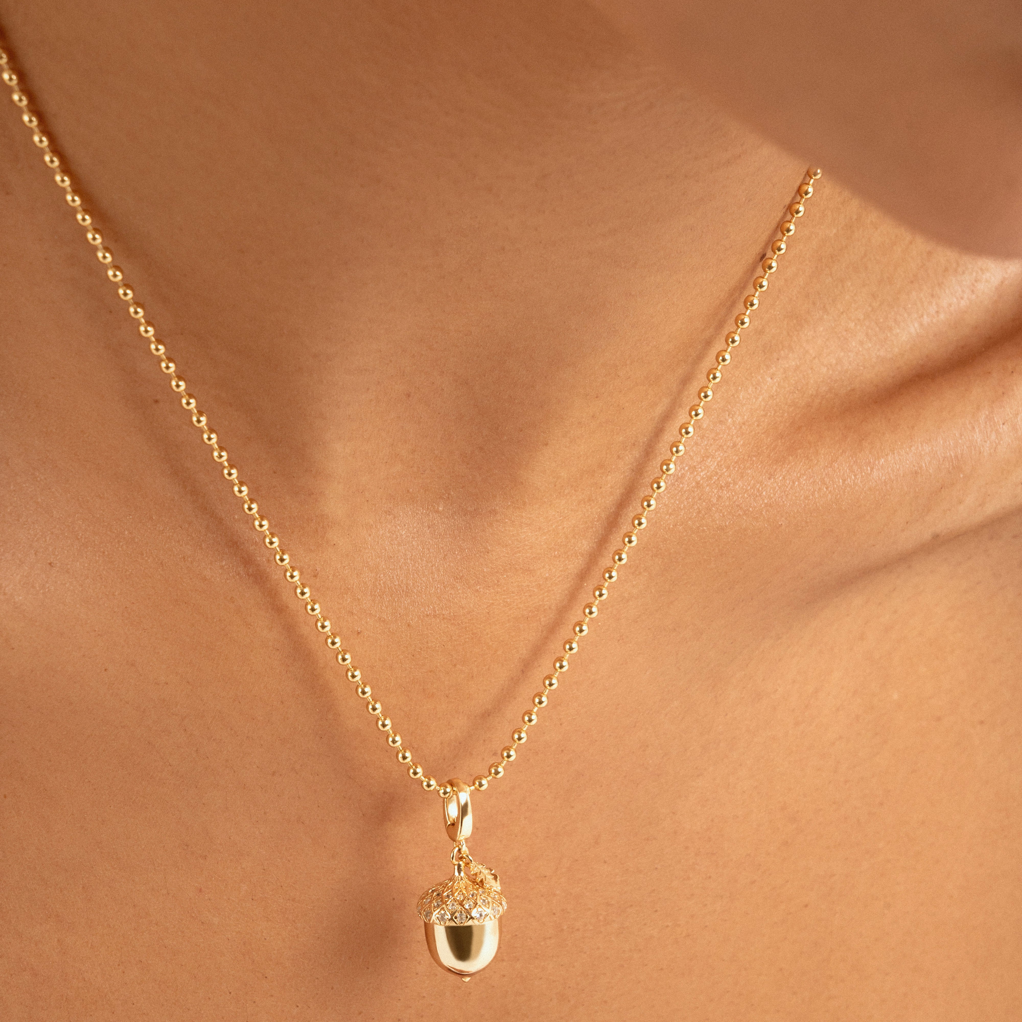 A close-up of a delicate gold chain necklace with an acorn pendant, worn on a model's neck.