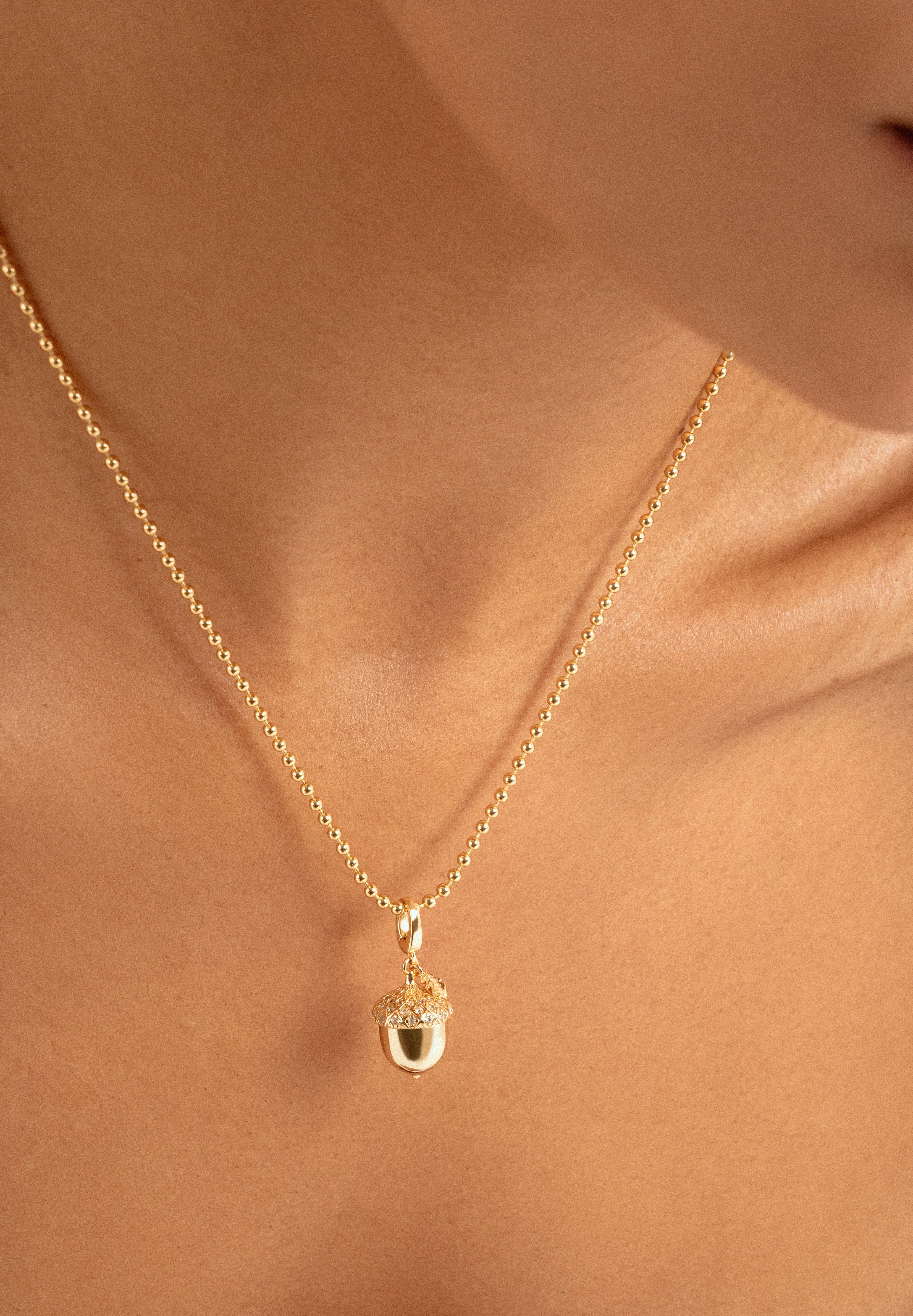 A close-up of a delicate gold chain necklace with an acorn pendant, worn on a model's neck.