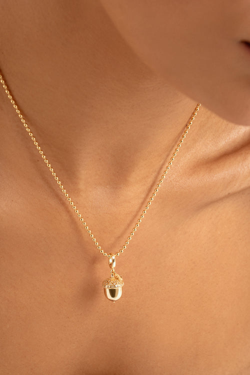 A close-up of a delicate gold chain necklace with an acorn pendant, worn on a model's neck.
