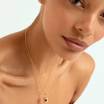 A close-up of a model wearing a delicate gold chain necklace with a small acorn shaped pendant set against a clean white background.