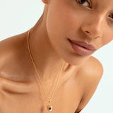 A close-up of a model wearing a delicate gold chain necklace with a small acorn shaped pendant set against a clean white background.
