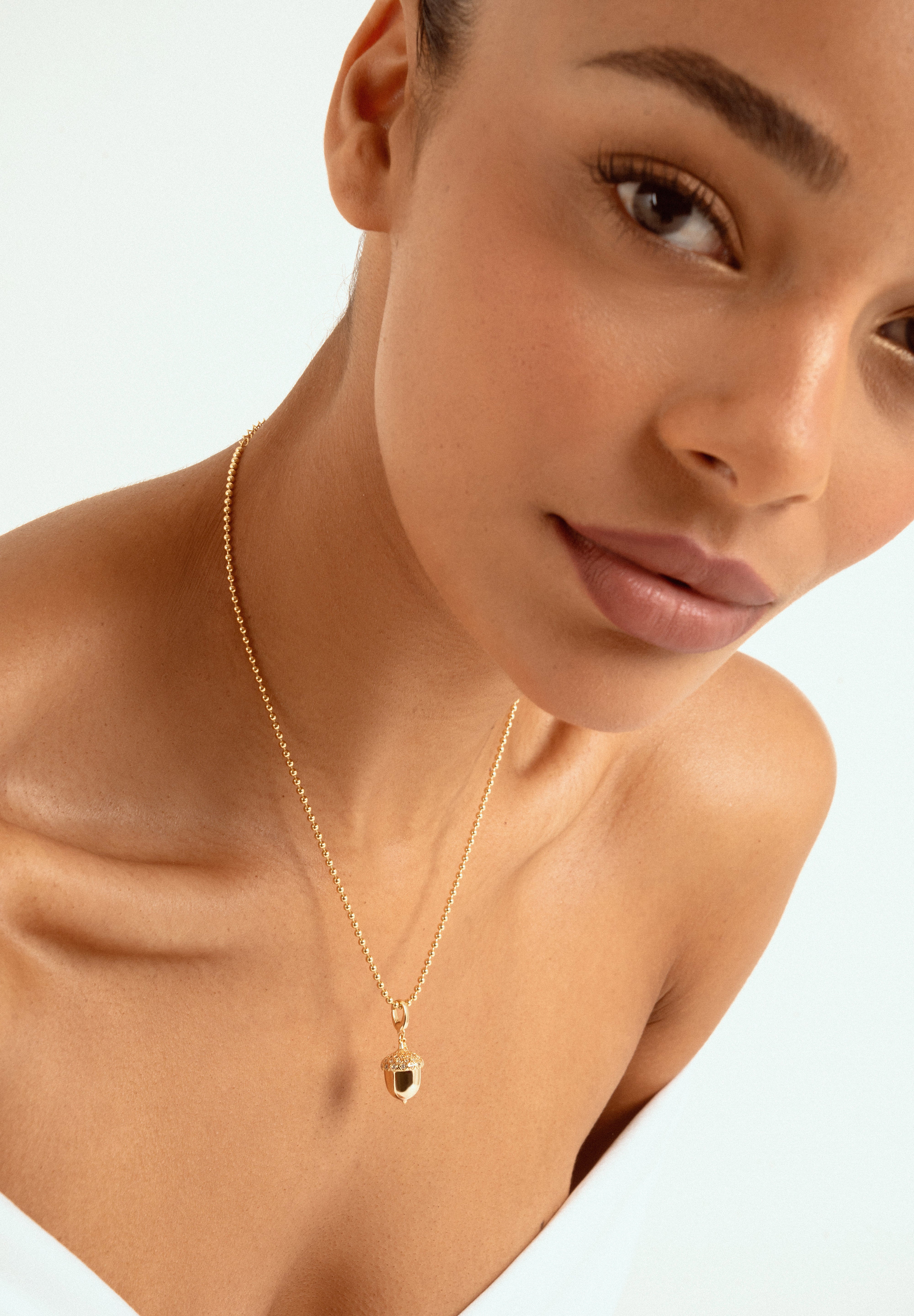 A close-up of a model wearing a delicate gold chain necklace with a small acorn shaped pendant set against a clean white background.