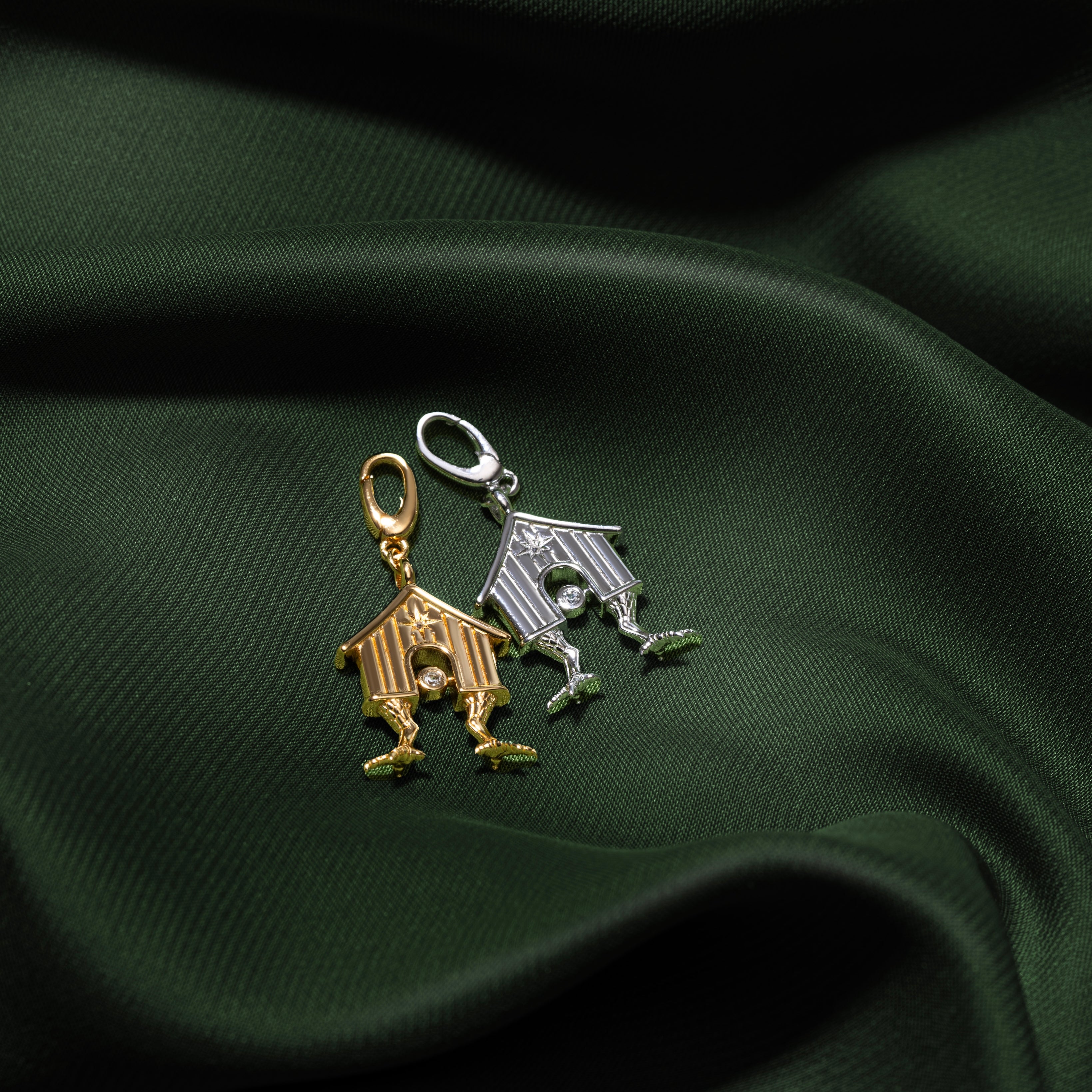 Two small house-shaped charms, one gold-toned and one silver-toned, displayed on draped dark green fabric.