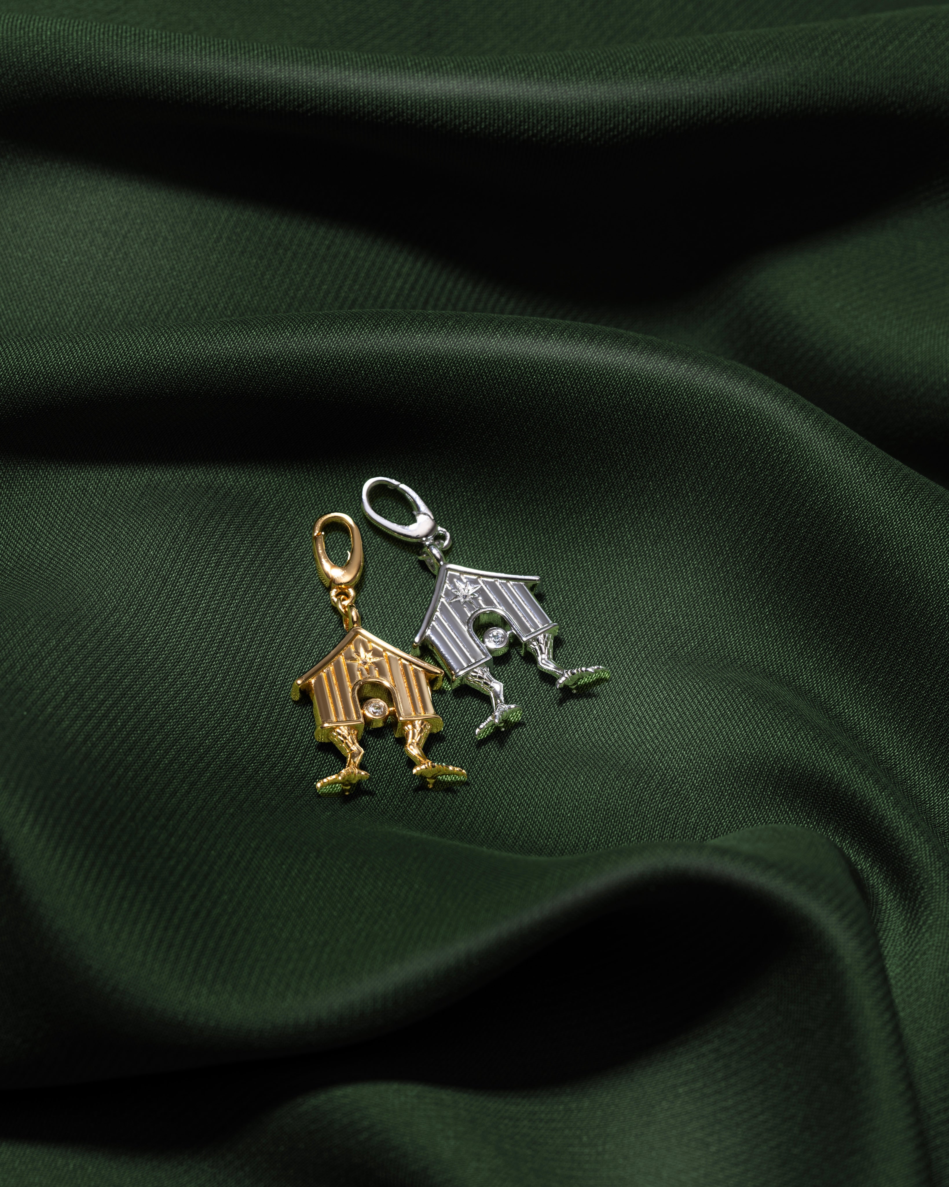 Two small house-shaped charms, one gold-toned and one silver-toned, displayed on draped dark green fabric.