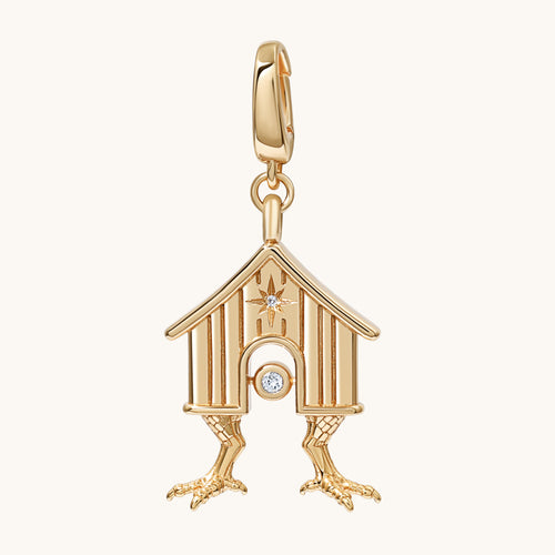 A gold house charm pendant with two legs and a small clear gemstone detail, shown on a light cream background.