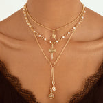A close-up of a model wearing a layered gold necklace set with pearl accents, a key pendant, and a long drop chain with moon and cross charms, styled with a black lace top.