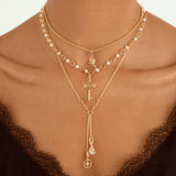 A close-up of a model wearing a layered gold necklace set with pearl accents, a key pendant, and a long drop chain with moon and cross charms, styled with a black lace top.
