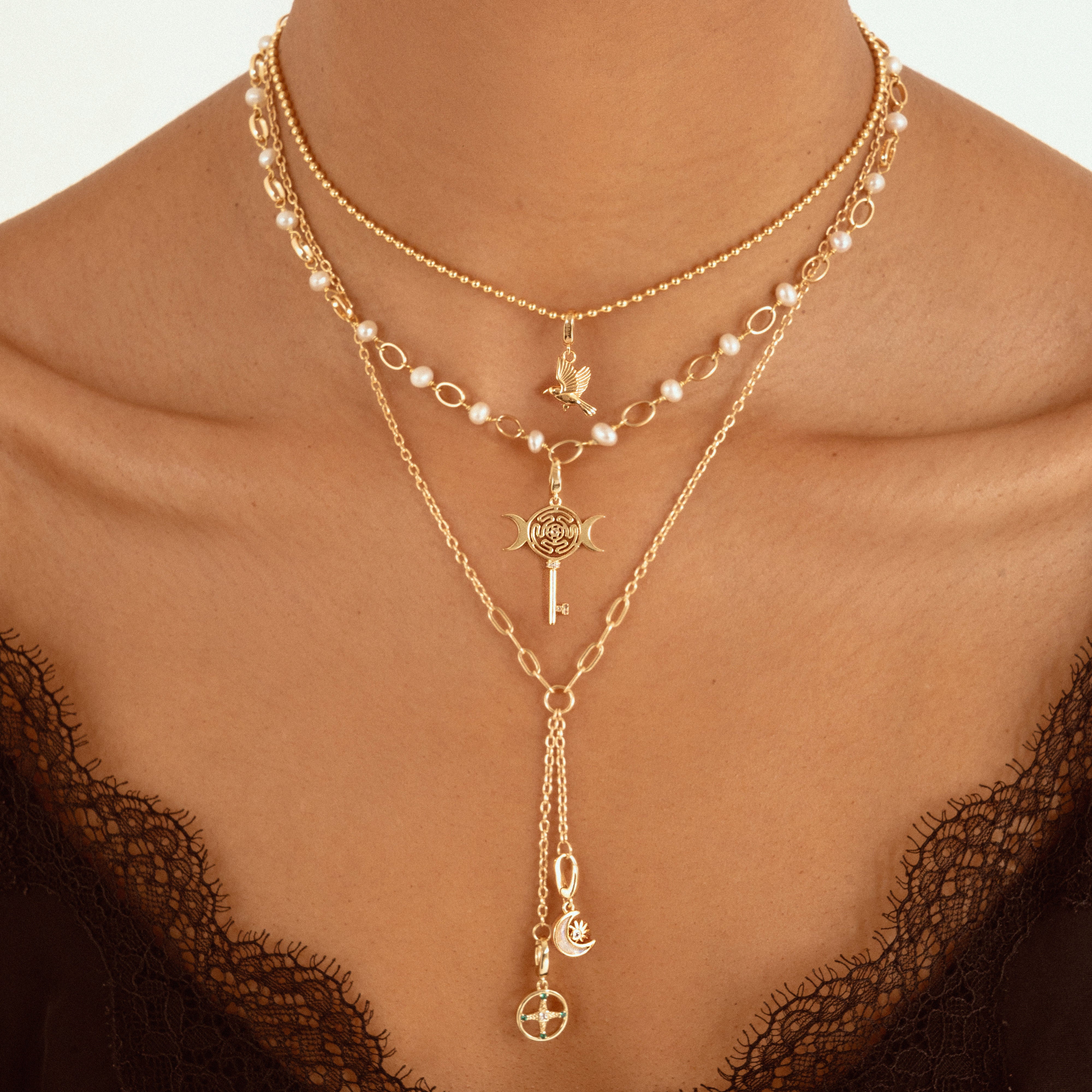 A close-up of a model wearing a layered gold necklace set with pearl accents, a key pendant, and a long drop chain with moon and cross charms, styled with a black lace top.