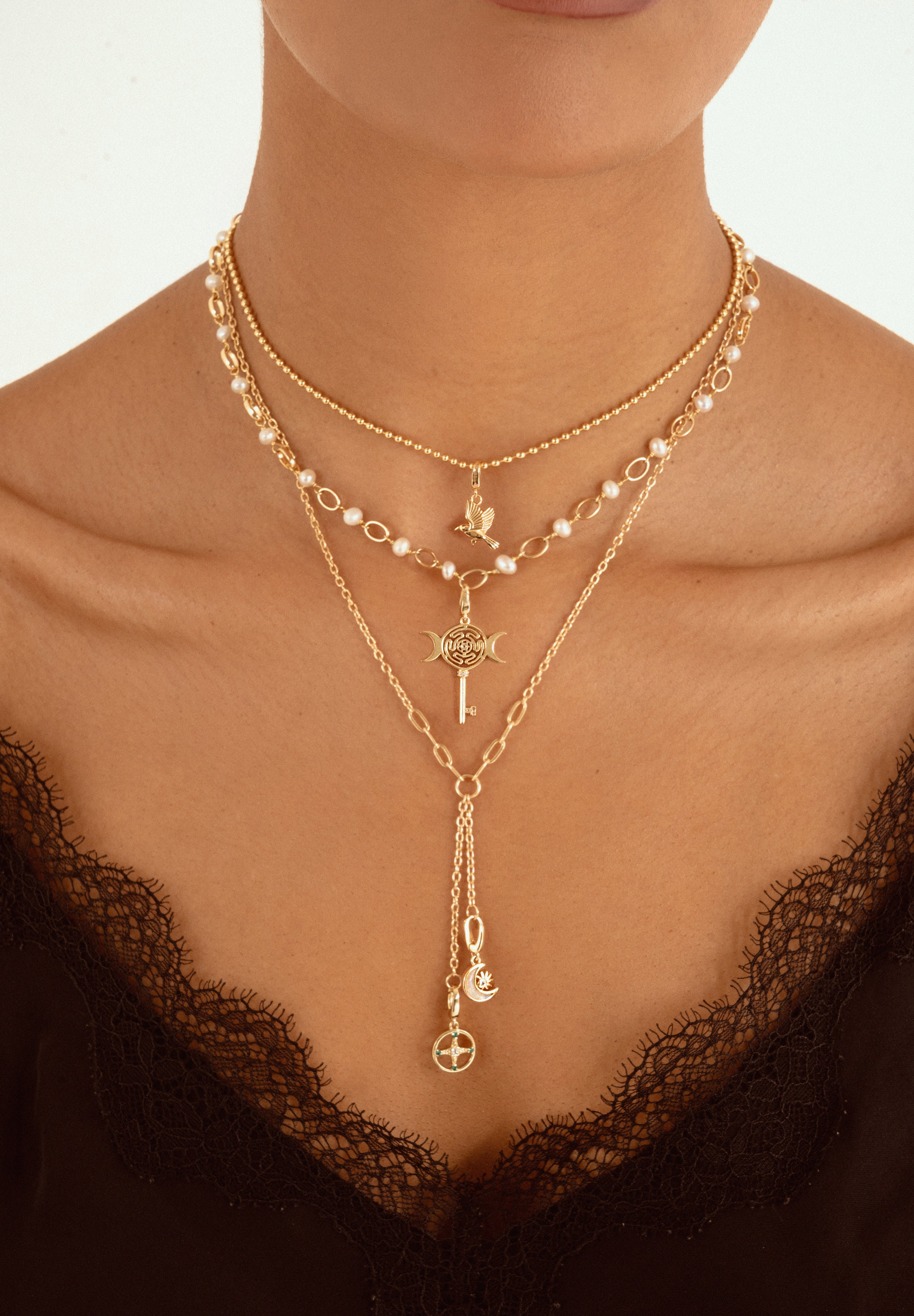 A close-up of a model wearing a layered gold necklace set with pearl accents, a key pendant, and a long drop chain with moon and cross charms, styled with a black lace top.