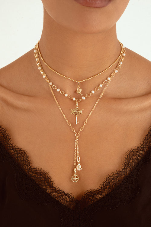 A close-up of a model wearing a layered gold necklace set with pearl accents, a key pendant, and a long drop chain with moon and cross charms, styled with a black lace top.