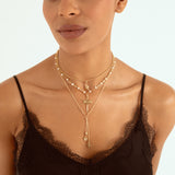 A close-up of a model wearing a layered gold necklace set with a cross pendant and pearl accents, styled over a black lace-trim camisole against a light background.