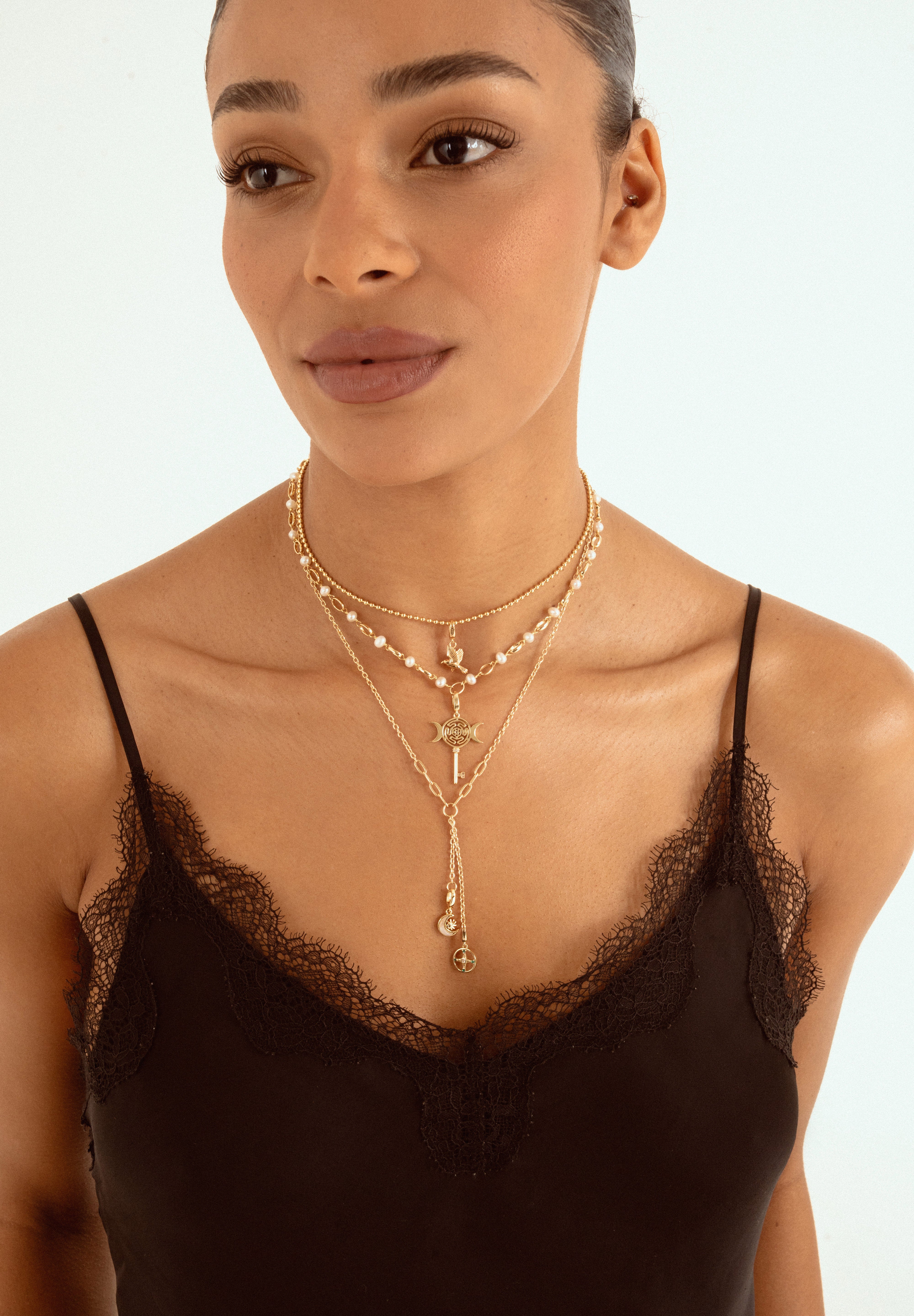 A close-up of a model wearing a layered gold necklace set with a cross pendant and pearl accents, styled over a black lace-trim camisole against a light background.