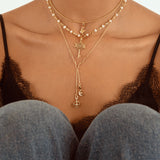 A close-up of a model wearing a layered gold necklace set with star, moon, and cross charms, styled over a black lace camisole and blue jeans.
