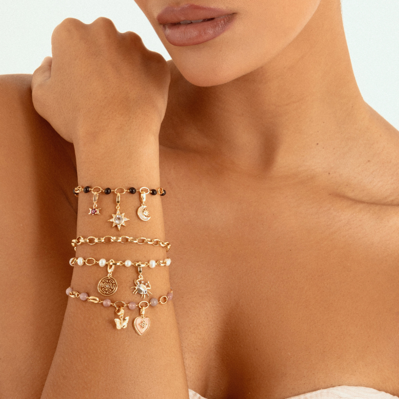 A close-up of a model's wrist wearing four delicate charm bracelets in gold-tone metal with beads and moon, star, and coin charms, against a clean white background.